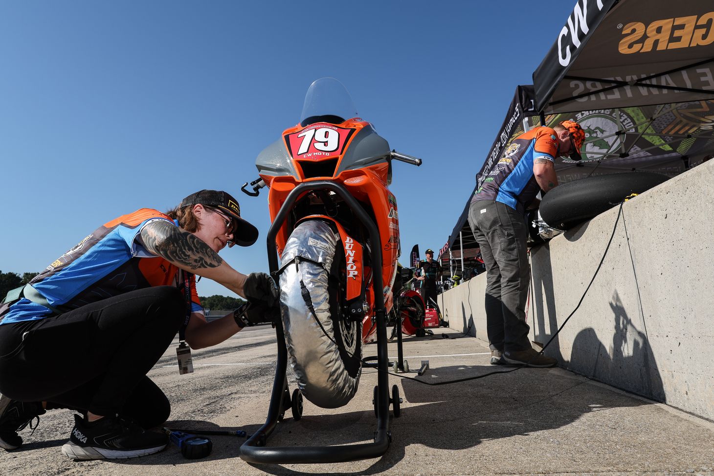 Sara is squatting down checking the front tire pressure on the orange and grey #79 CW Moto Racing Yamaha R1.